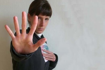 Eine Frau blickt leicht nach unten in die Kamera, hält in einer Hand mehrere Geldscheine an ihre Brust und streckt den Arm und die Handfläche der anderen Hand vor ihrem Oberkörper wie als Stoppsignal aus.