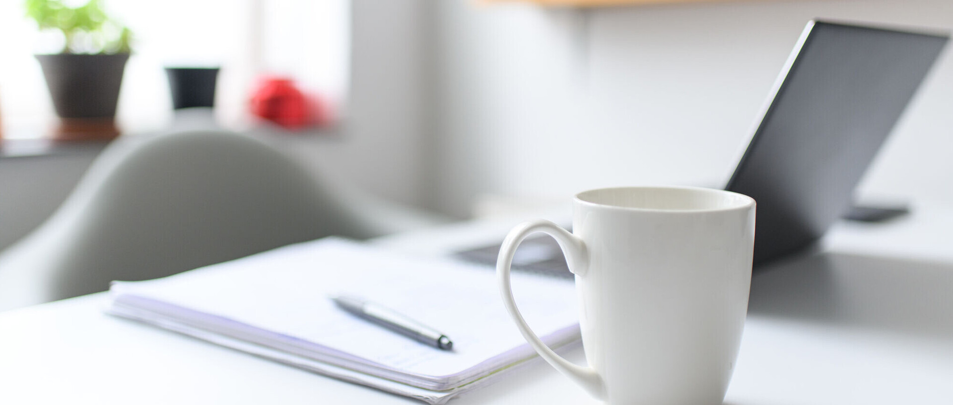 Eine Tasse steht auf einem Tisch. Im Hintergrund liegen ein Notizblock und ein Laptop.