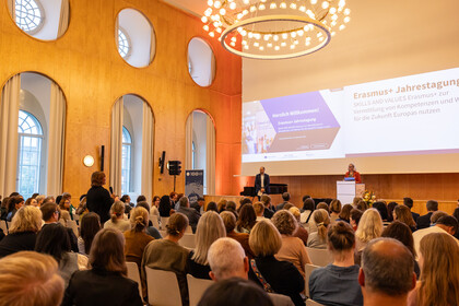 Zwei Personen stehen auf der Bühne der Aula. Eine Person aus dem Publikum ist aufgestanden, um eine Frage zu stellen.