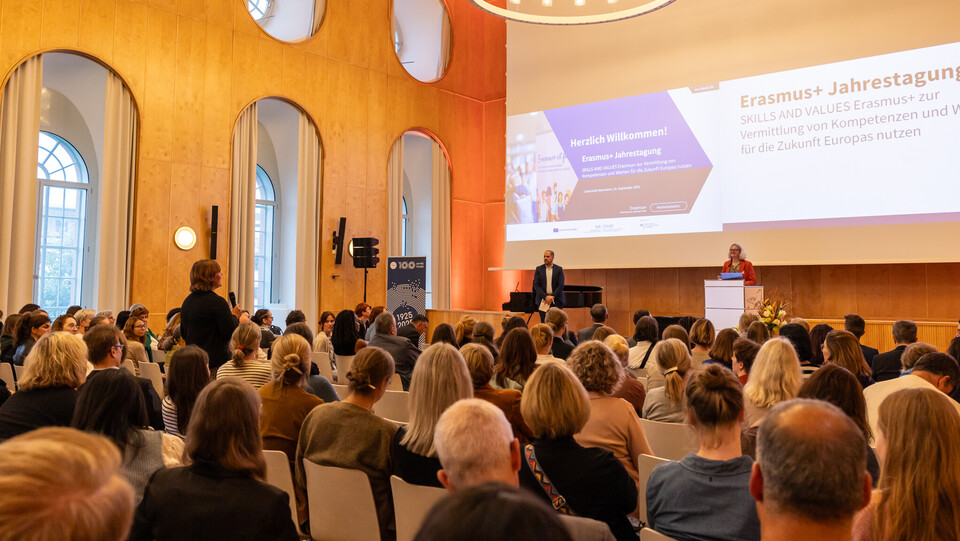 Zwei Personen stehen auf der Bühne der Aula. Eine Person aus dem Publikum ist aufgestanden, um eine Frage zu stellen.