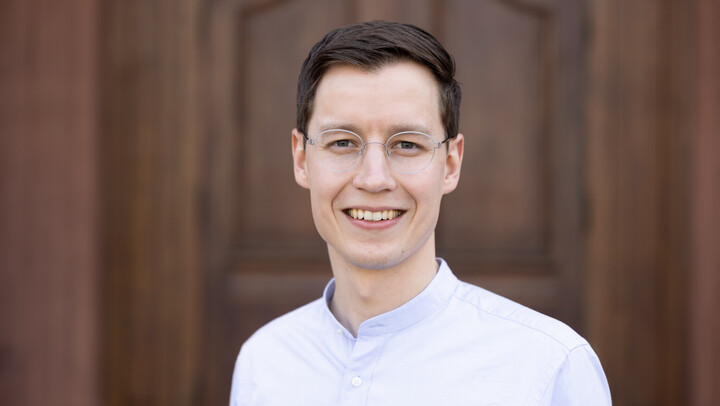 Kai Maeckle. He is wearing a white sweater and is standing in front of a brown door.