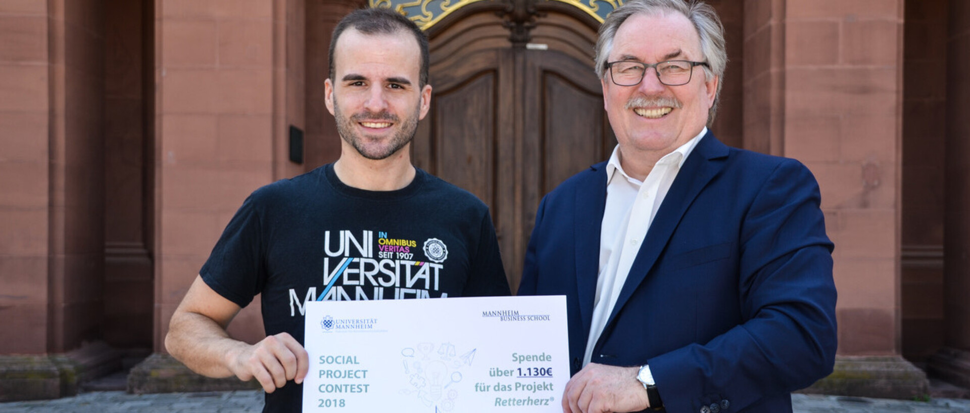Ein Mann im Anzug und ein Mann im schwarzen T-Shirt mit der Aufschrift "Universität Mannheim" stehen im Ehrenhof des Schlosses und halten ein Spendenschild in der Hand.