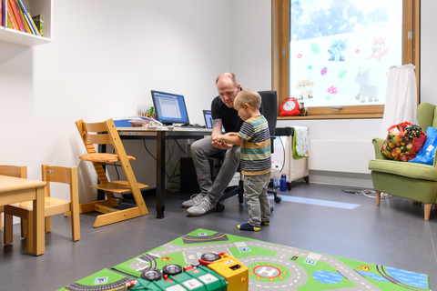Vater und Sohn sitzen in einem Spielraum, der Vater zeigt dem Sohn etwas.