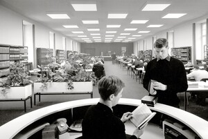 schwarz-weiß Aufnahme des damaligen Lesesaals der Universität Mannheim. Ein männlich gelesener Student steht vor der Theke und hält ein Buch in der Hand. Die Person hinter der Theke liest ein Buch. 