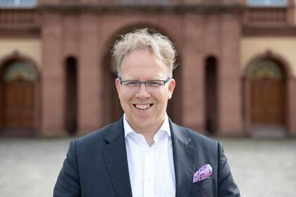 Portrait von Oliver Brand. Er trägt ein weißes Hemd sowie einen schwarzen Blazer und steht vor dem Mannheimer Schloß.