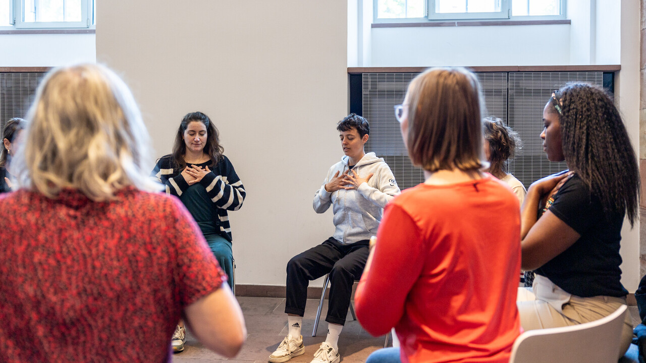 Personen im Stuhlkreis in den Katakomben der Universität. Alle haben die Augen geschlossen und beide Hände auf die Brust gelegt.