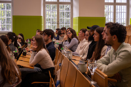Eine Gruppe Studierender sitzt in einem Hörsaal an Tischreihen, sie sind von der Seite zu sehen. 
