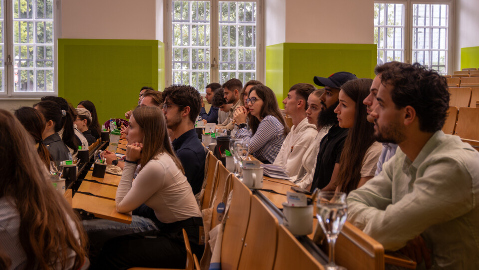 Eine Gruppe Studierender sitzt in einem Hörsaal an Tischreihen, sie sind von der Seite zu sehen.