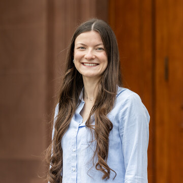 Evelyn Bentz hat lange, dunkelbraune, lockige Haare, trägt eine hellblaue Bluse und lächelt freundlich in die Kamera.