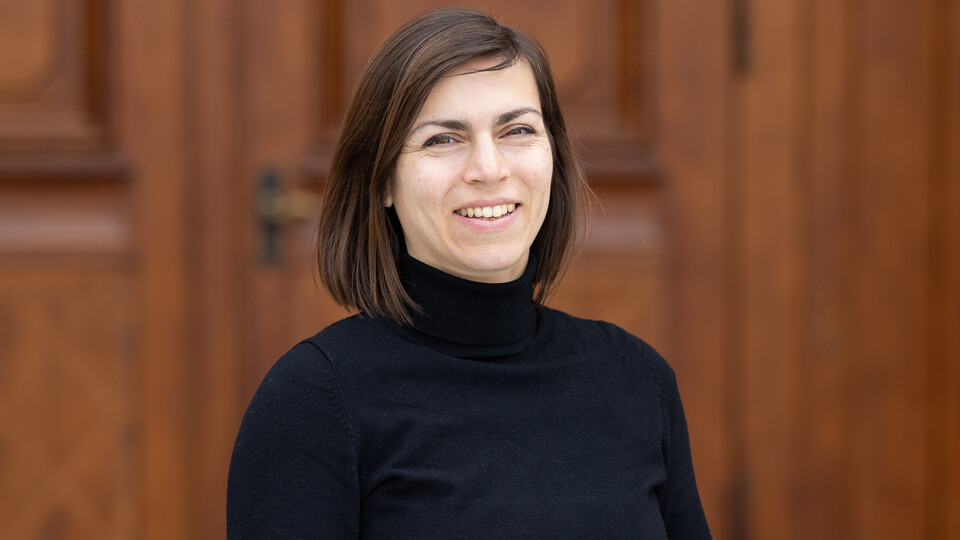 Porträt von Anna-Sophie Kurella, die lächelnd und mit schwarzem Rollkragenpullover vor einer Holztür steht.