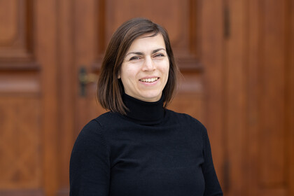 Porträt von Anna-Sophie Kurella, die lächelnd und mit schwarzem Rollkragenpullover vor einer Holztür steht.