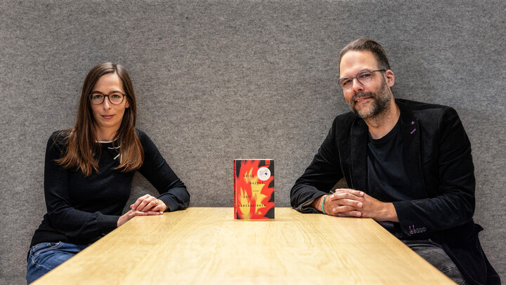 Sandra Beck und Thomas Wortmann sitzen an einem hellen Holztisch vor einer grauen Wand. Auf dem Tisch zwischen ihnen ist ein Buch mit gelb-rotem Cover aufgestellt. Es ist der Roman "Drei Kameradinnen" von Shida Bazyar.