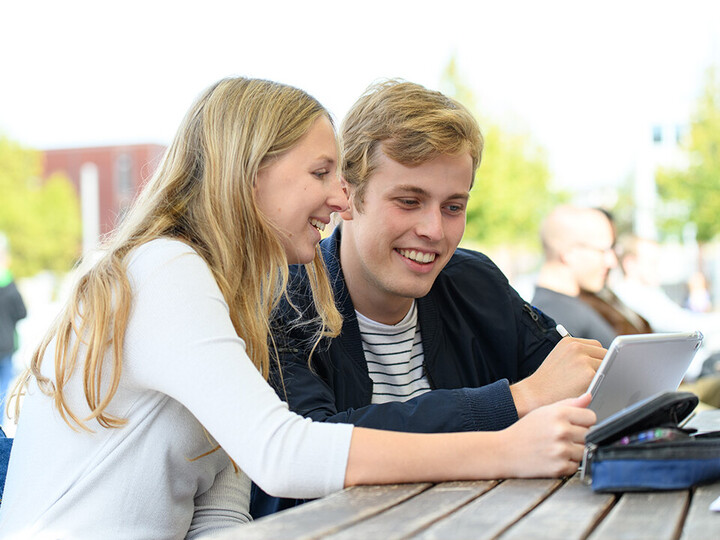 Zwei Studierende sitzen lachend über ein Ipad gebeugt draußen an einem Tisch.