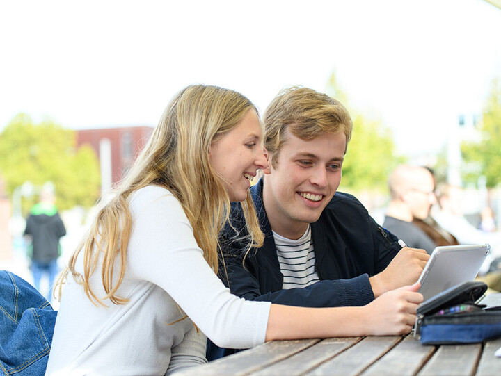 Zwei Studierende sitzen lachend über ein Ipad gebeugt draußen an einem Tisch.