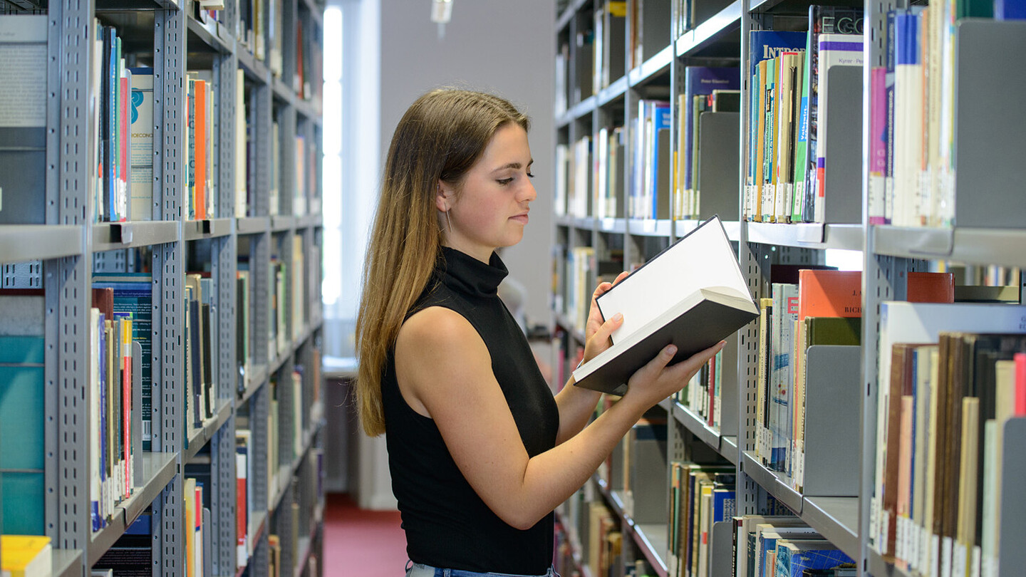 Eine Studentin steht vor einem Bibliotheksregal und liest in einem Buch.