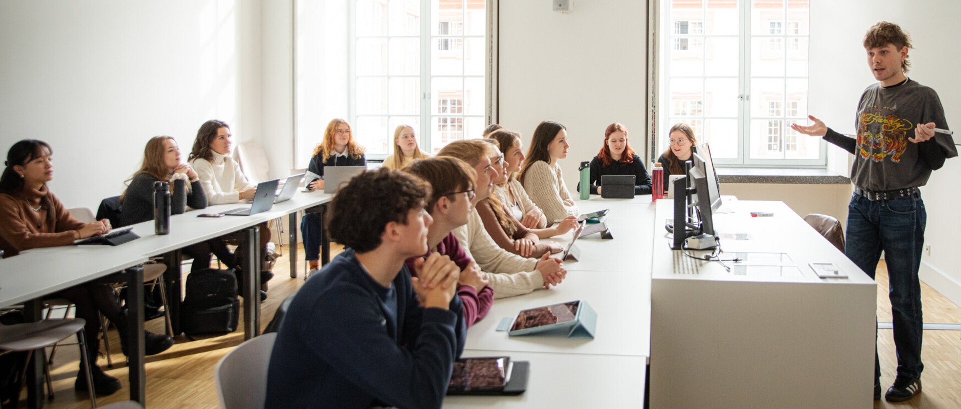Eine Lehrperson steht an der Tafel. Die Person gestikuliert und spricht zu der Gruppe von 14 Studierenden, die im Semiarraum an Tischen sitzen.
