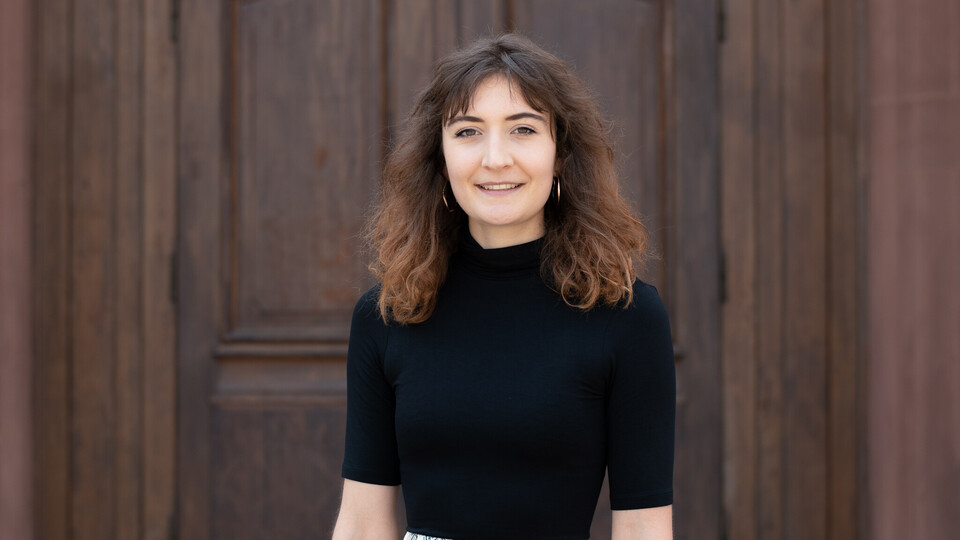 Portrait of Jessica Müller. She is standing in front of a door, waering a black shirt.