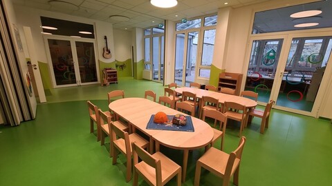 Ein freundlicher und heller Kindergartenraum mit grünem Boden und bunten Möbeln. Um zwei ovale Holztische sind Kinderstühle gruppiert, im Hintergrund befinden sich eine Gitarre, die an der Wand hängt, sowie eine Tür in einen anderen Spielbereich mit bunten Kreisen an den Fenstern.