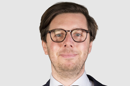 Portrait von Philipp Dörrenberg. Er trägt einen dunkelblauen Anzug mit brauner Krawatte sowie eine Brille.