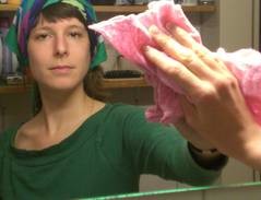 Eine Frau mit einem bunten Haarband und in einem grünen Oberteil putzt mit einem rosafarbenen Tuch den Spiegel, in dem sie sich selbst sieht.