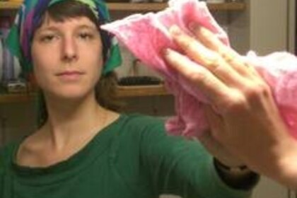 Eine Frau mit einem bunten Haarband und in einem grünen Oberteil putzt mit einem rosafarbenen Tuch den Spiegel, in dem sie sich selbst sieht.