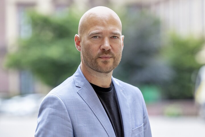 Herr Traunmüller hat eine Glatze, er trägt ein schwarzes T-Shirt und ein hellblaues Jacket. Er steht im Freien vor einem Baum