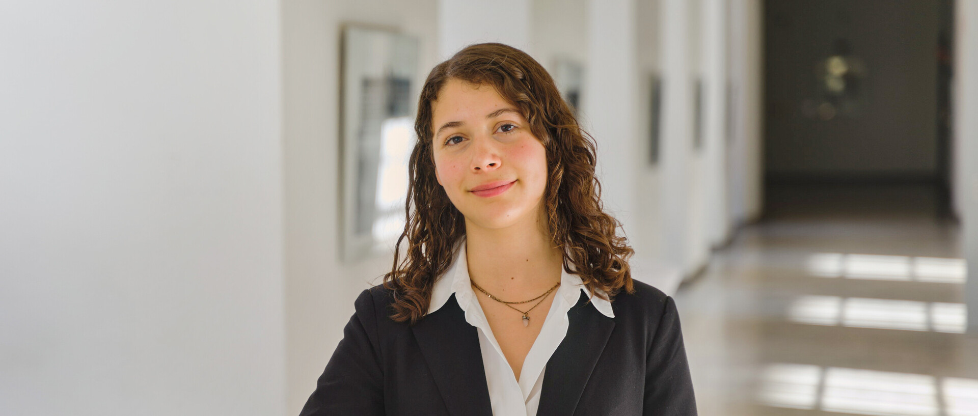 Porträt von Emiliana. Sie steht in einem Gang der Universität, trägt eine weiße Bluse und ein schwarzes Jacket und schaut freundlich in die Kamera.