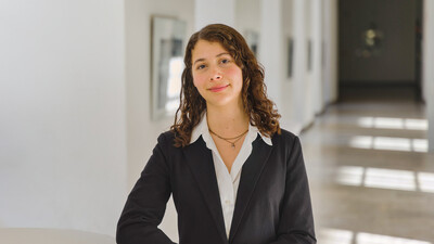 Porträt von Emiliana. Sie steht in einem Gang der Universität, trägt eine weiße Bluse und ein schwarzes Jacket und schaut freundlich in die Kamera.