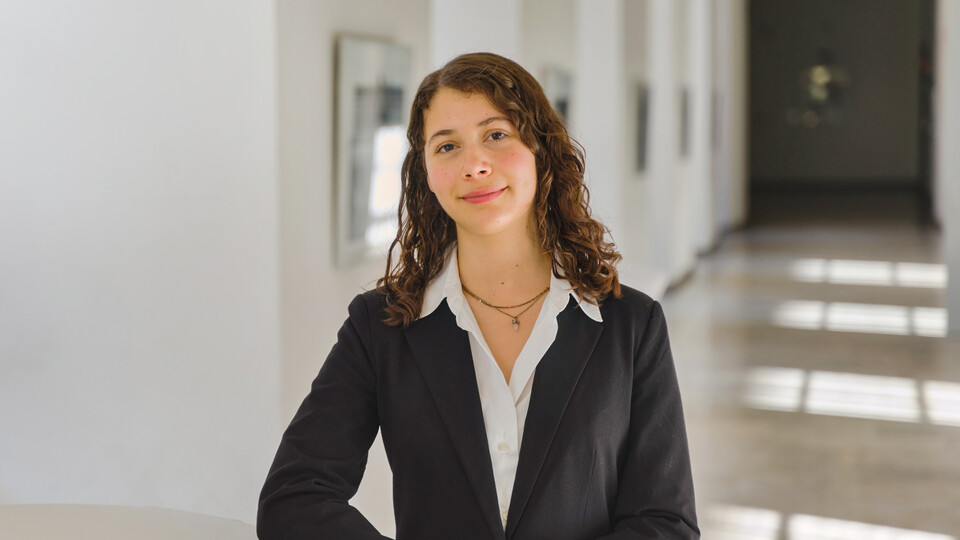 Porträt von Emiliana. Sie steht in einem Gang der Universität, trägt eine weiße Bluse und ein schwarzes Jacket und schaut freundlich in die Kamera.