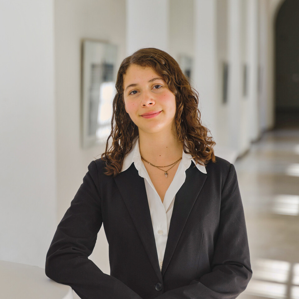 Porträt von Emiliana. Sie steht in einem Gang der Universität, trägt eine weiße Bluse und ein schwarzes Jacket und schaut freundlich in die Kamera.