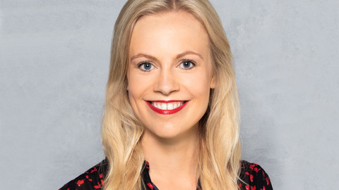 Inga Schulz. She is wearing a black and red blouse and is standing in front of grey wall.