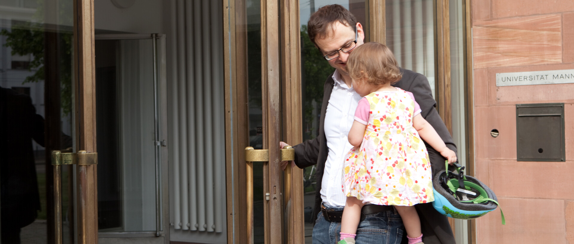 Ein Vater hält sein Kind auf dem Arm und lächelt. Gleichzeitig öffnet er eine Eingangstür zum Schloss Mannheim.