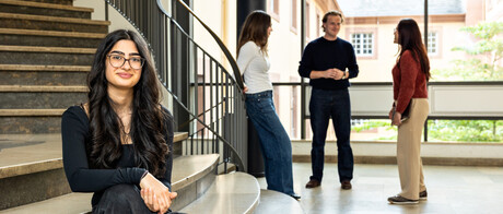 Eine Studentin sitzt auf der Treppe im Treppenhaus des Schlosses. Im Hintergrund unterhalten sich drei Studierende. 