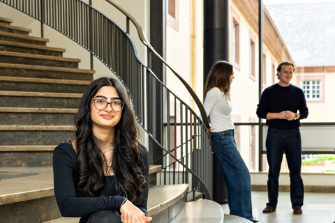 Eine Studentin sitzt auf der Treppe im Treppenhaus des Schlosses. Im Hintergrund unterhalten sich drei Studierende. 