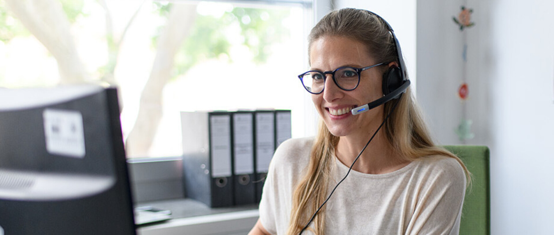Eine Person sitzt lächelnd an einem Schreibtisch. Sie blickt auf einen Computerbildschirm und trägt ein Headset.