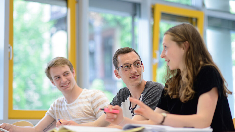 Drei Studierende unterhalten sich in einem Seminarraum.