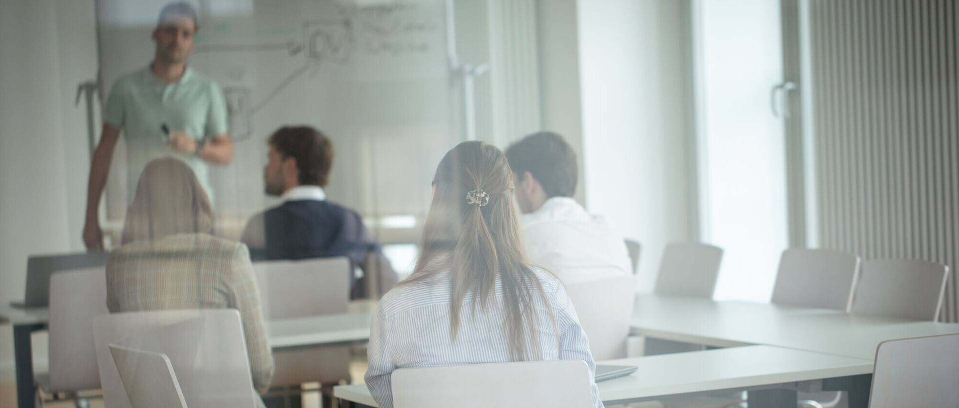 Vier Personen sitzen in einem modern ausgestatteten Seminarraum. Ein junger Mann steht vor dem Whiteboard.