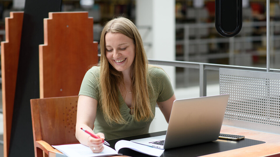 Eine Studentin sitzt an einem Tisch mit einem aufgeklapptenn Laptop und einem Stift in der Hand.