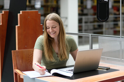 Eine Studentin sitzt an einem Tisch mit einem aufgeklapptenn Laptop und einem Stift in der Hand.