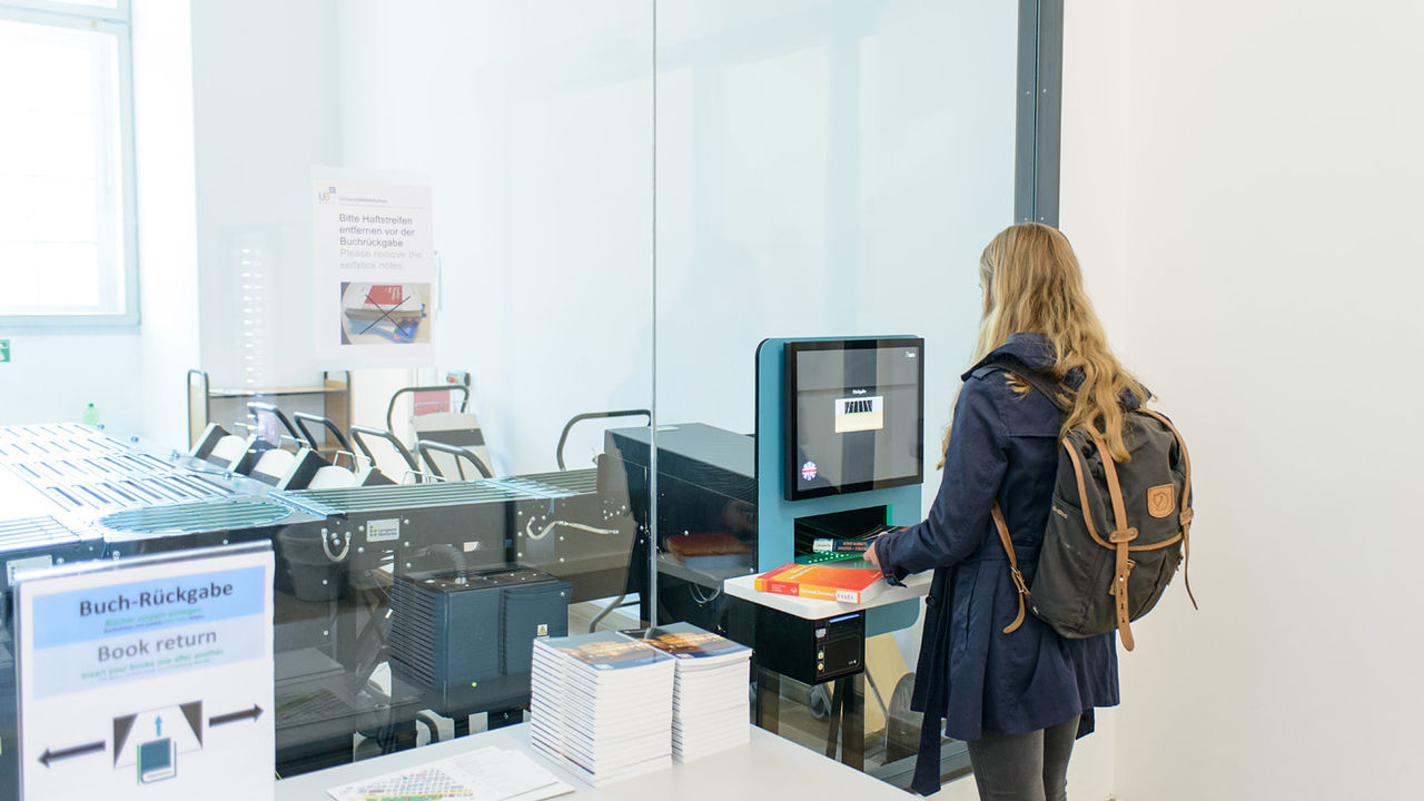Eine Studentin gibt Bücher an der Rückgabeanlage des Ausleihzentrums der Universität Mannheim zurück. Sie trägt blondes welliges Haar, eine dunkelblaue Jacke und einen Rucksack.