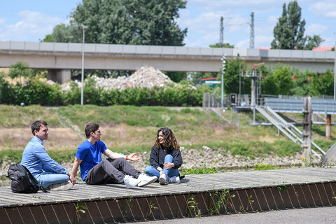Three students sit in the sun on a wooden walkway. A bridge can be seen in the background.