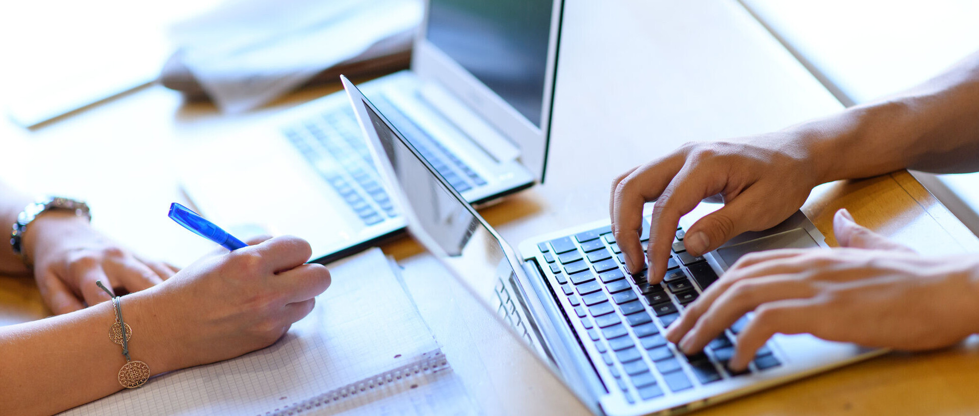 Zwei Laptops stehen versetzt auf einem Tisch. Am rechten Laptop sind zwei Hände an der Tastatur. Gegenüber davon schreibt eine Person auf einen Block.
