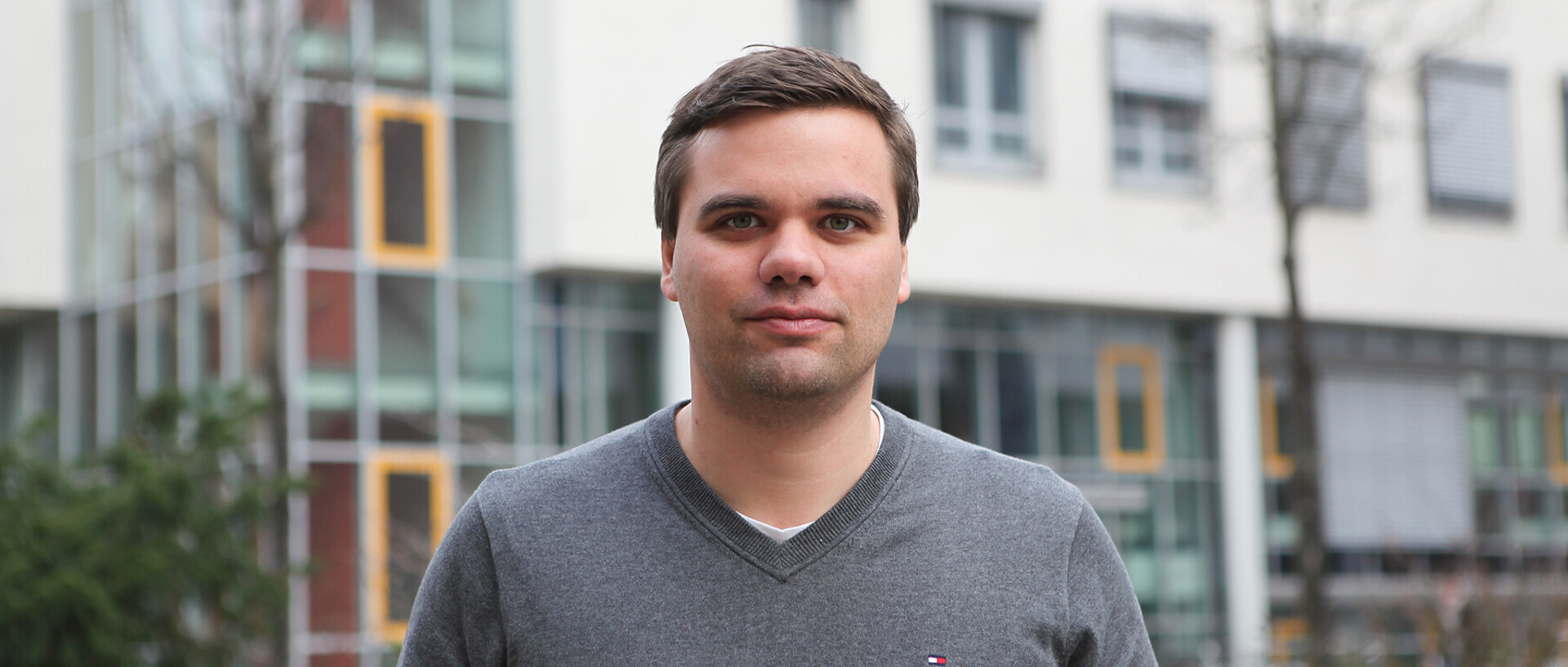 Personenportrait eines Mannes mit einem grauen Pullover vor einem Gebäude. 