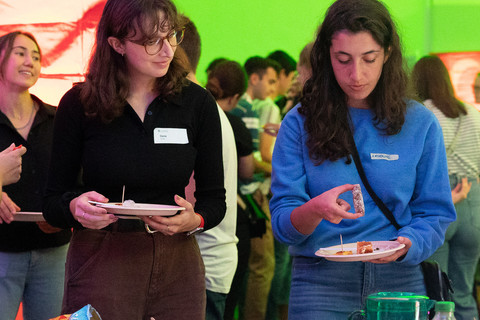 Zwei Studentinnen stehen vor einem Tisch mit Essen. Beide halten einen Teller in der Hand.