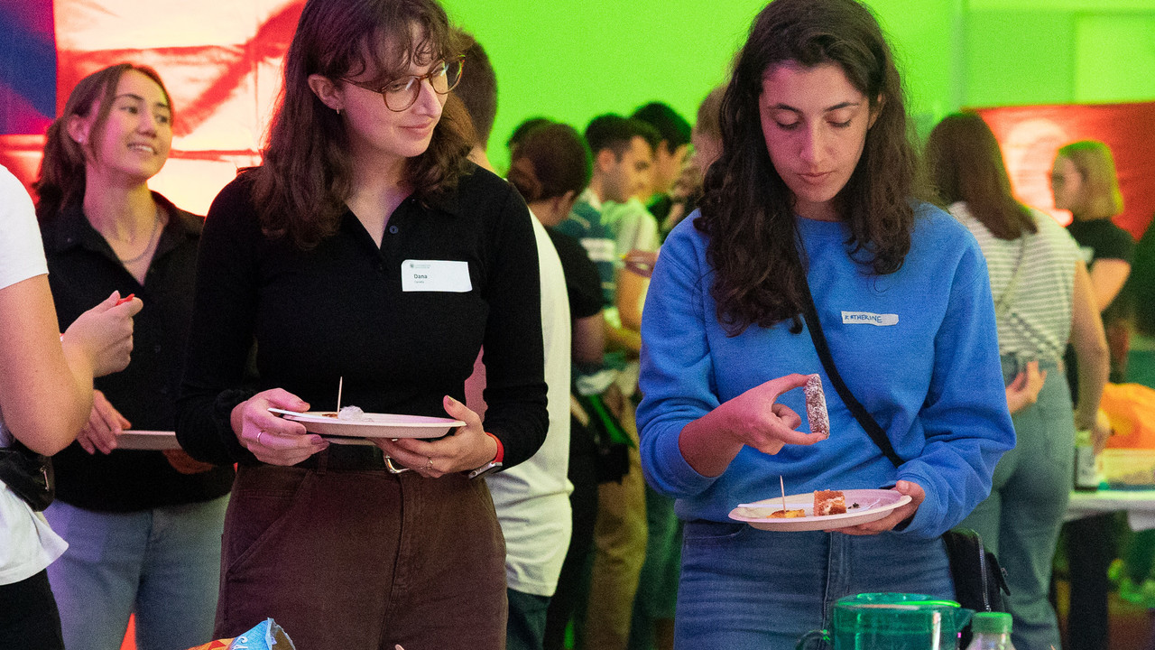 Zwei Studentinnen stehen vor einem Tisch mit Essen. Beide halten einen Teller in der Hand.