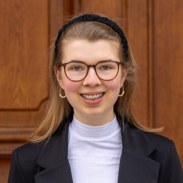 Jolina Sieland. Jolina trägt Brille, einen Haarreif und einen schwarzen Blazer. Sie lächelt freundlich in die Kamera.