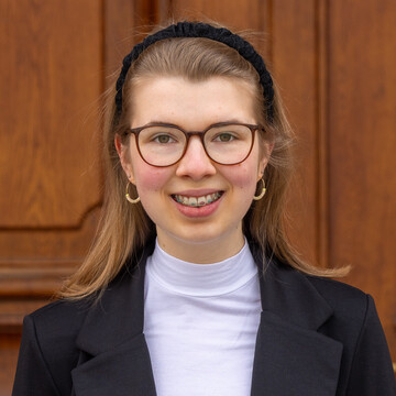Jolina Sieland. Jolina trägt Brille, einen Haarreif und einen schwarzen Blazer. Sie lächelt freundlich in die Kamera.