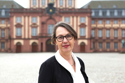 Portrait von Sabine Sonnentag. Sie trägt eine weiße Bluse und einen schwarzen Blazer. Sie steht vor dem Schloss auf dem Ehrenhof.