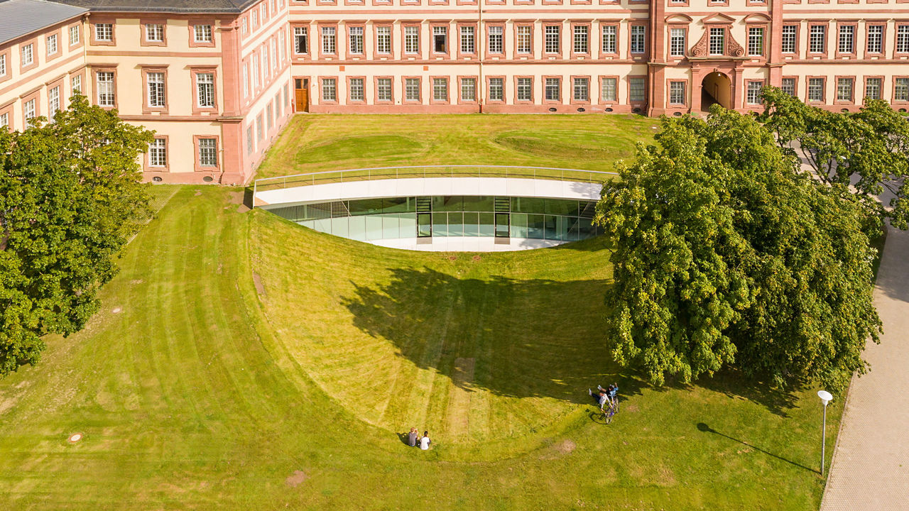 Die mit grünem Rasen überwachsenen Räume der Mannheim Business School. Die Räume sind im Souterrain vor dem Schloss und haben eine große Glasfront.