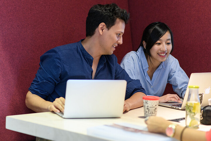 Studierende sitzen gemeinsam an einem Tisch und blicken lächelnd in einen Laptop.